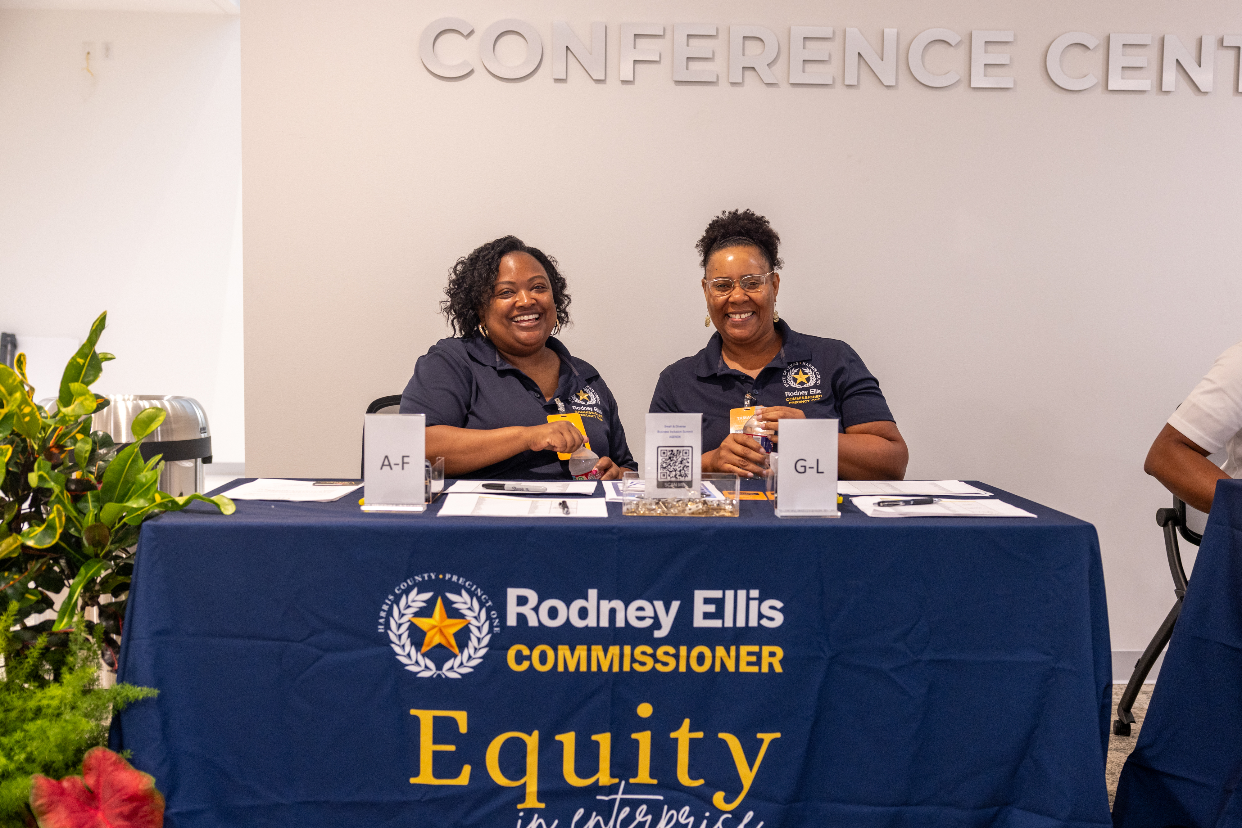 Two smiling registration desk volunteers behind a Rodney Ellis Commissioner Equity in Enterprise banner